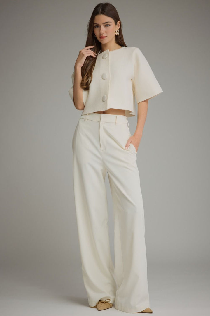 A woman modeling a cream-colored two-piece outfit consisting of a short-sleeve cropped jacket with three front buttons and matching high-waisted wide-leg trousers, set against a plain gray studio background.