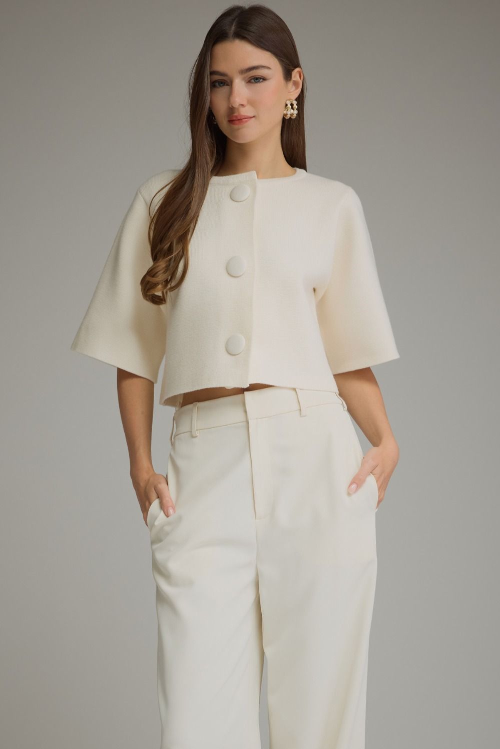 A woman modeling a cream-colored cropped jacket with three large decorative buttons down the front, paired with matching cream wide-leg trousers, against a solid gray studio background.