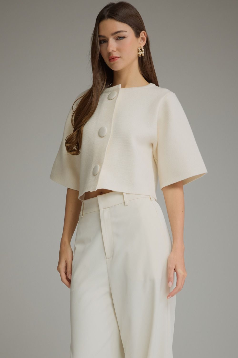 A woman modeling a cream-colored two-piece outfit consisting of a short-sleeve cropped jacket with large decorative buttons and matching high-waisted trousers, set against a plain gray studio background.