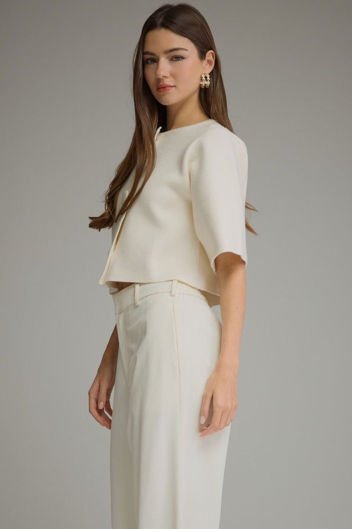 A woman modeling a cream-colored two-piece outfit consisting of a short-sleeve cropped jacket and matching high-waisted trousers, set against a plain gray studio background.