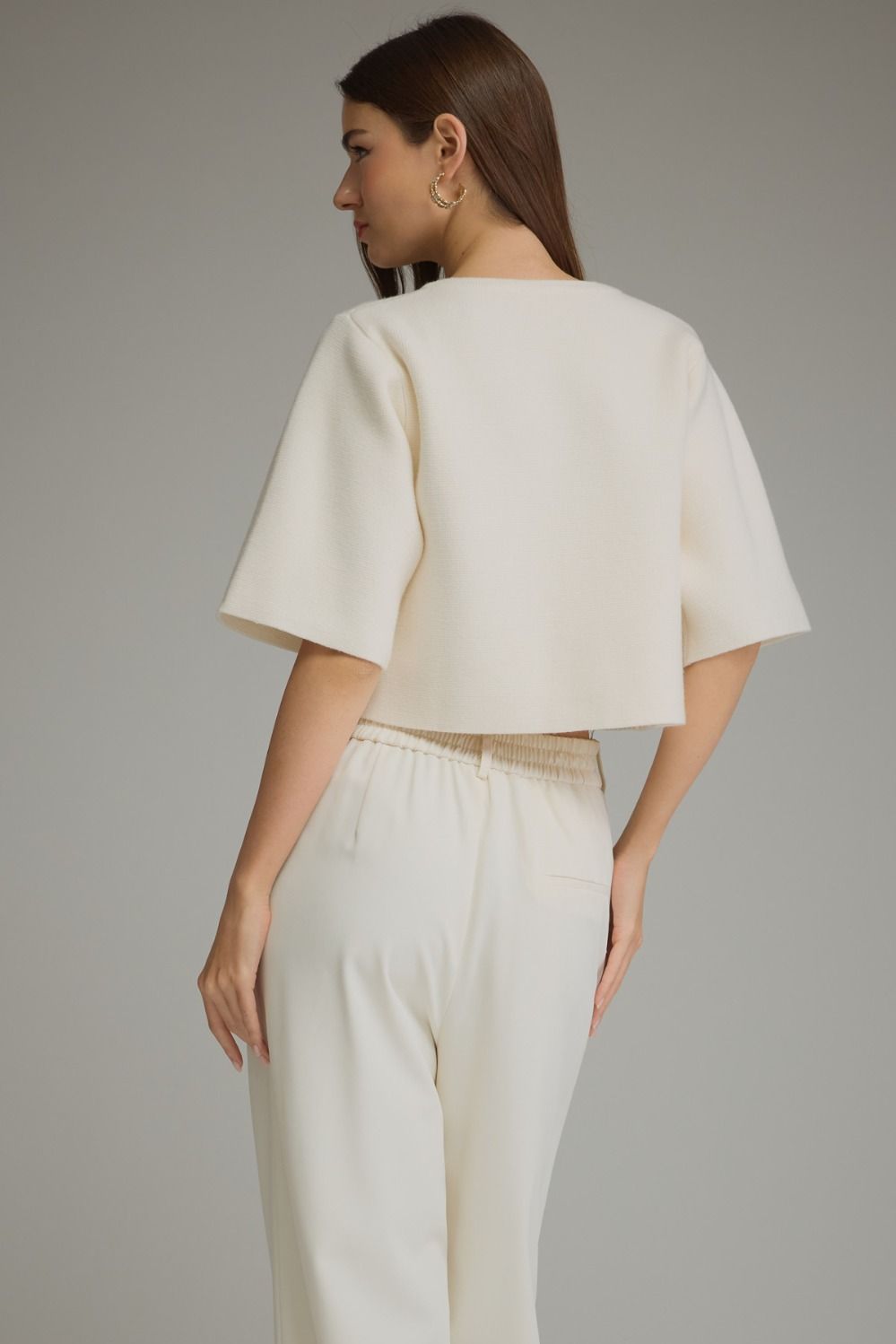 A woman shown from the back wearing a cream-colored short-sleeve cropped top and matching cream trousers against a plain gray studio background.