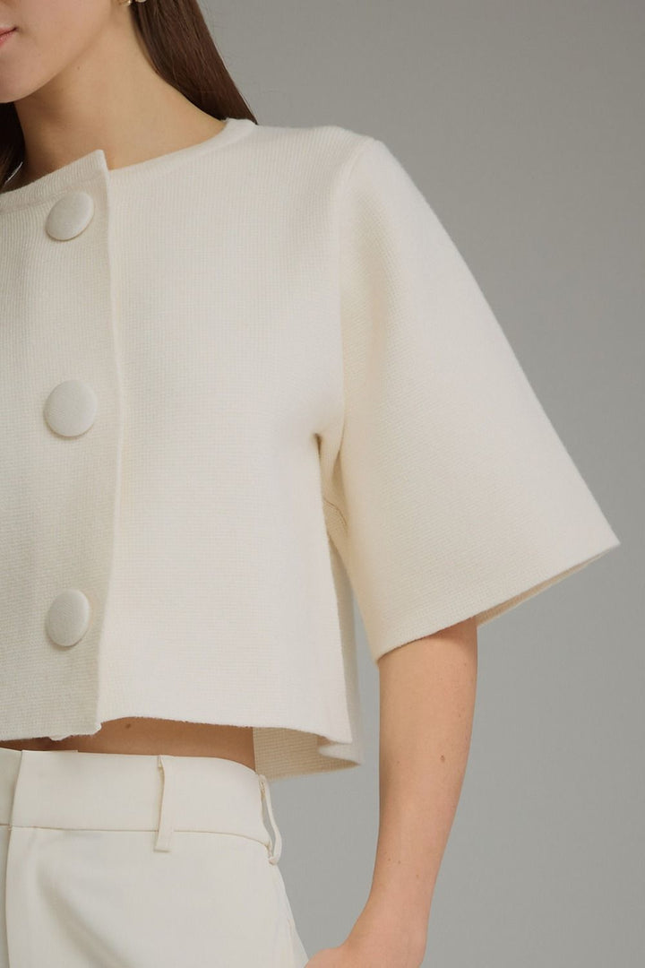 A close-up of a model wearing a cream-colored cropped jacket with wide short sleeves and three large decorative buttons down the front, paired with matching cream trousers against a neutral gray background.