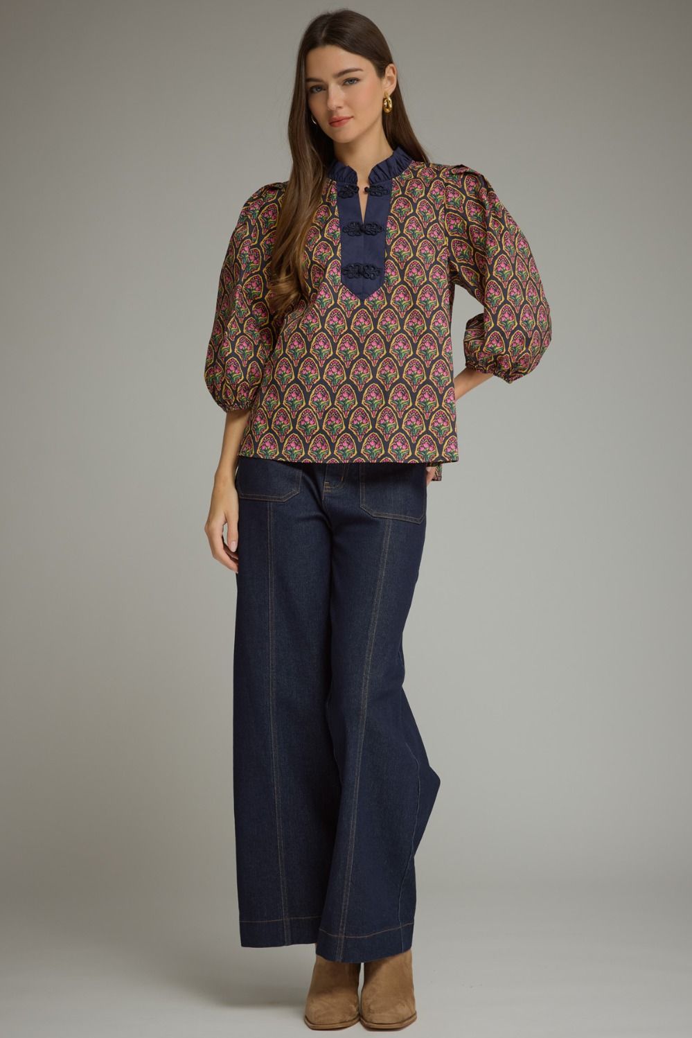A woman modeling a patterned long-sleeve blouse with a navy blue collar detail, paired with dark blue wide-leg jeans and tan ankle boots, against a neutral gray studio background.