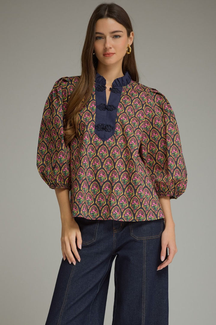 A woman modeling a patterned blouse with puff sleeves and a navy blue collar detail, paired with dark blue jeans, against a solid gray studio background.