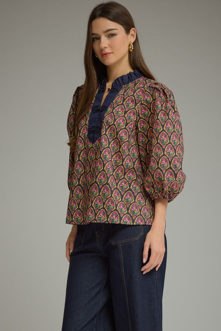 A woman modeling a long-sleeve blouse with a dense paisley pattern in pink, green, and gold over a dark base, featuring a dark blue ruffled split neckline. She is wearing dark blue jeans against a solid gray studio background.