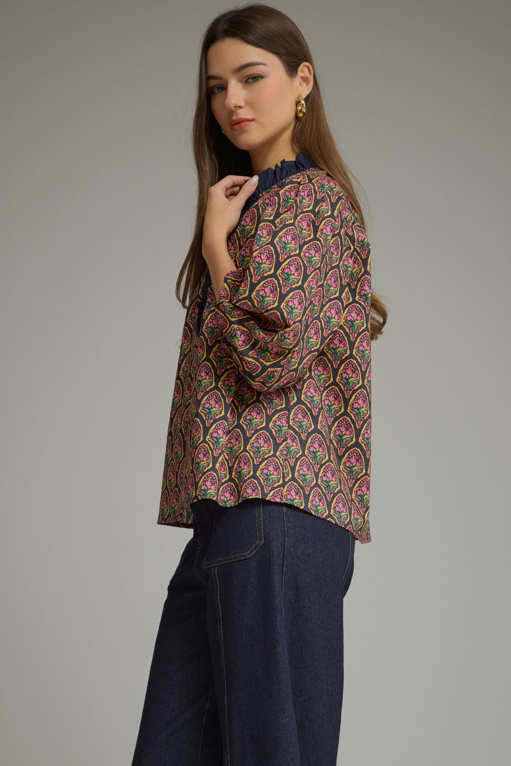 A woman modeling a long-sleeve blouse with an intricate multicolor paisley pattern in pink, green, and gold on a dark base, paired with dark blue jeans, against a solid gray studio background.