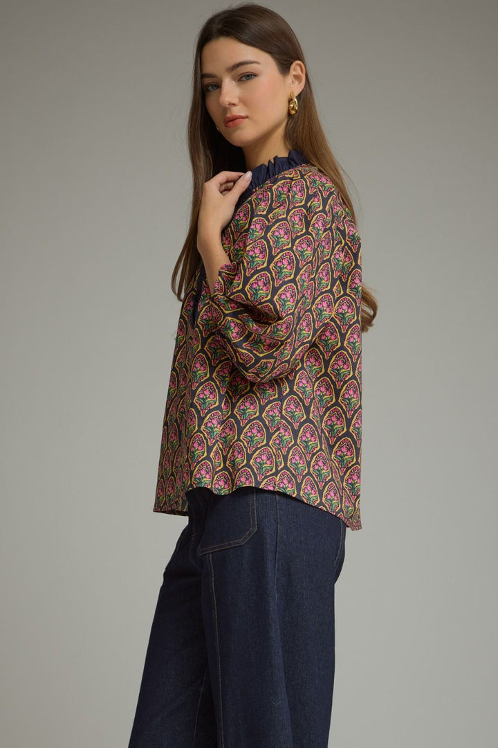 A woman modeling a long-sleeve blouse with an intricate multicolor paisley pattern in pink, green, and gold on a dark base, paired with dark blue jeans, against a solid gray studio background.
