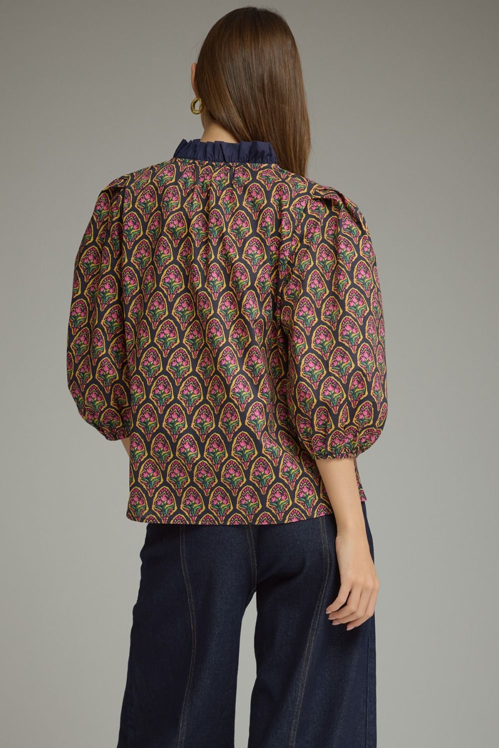 A woman shown from the back wearing a patterned blouse with puff sleeves and dark blue jeans, photographed against a plain gray studio background.