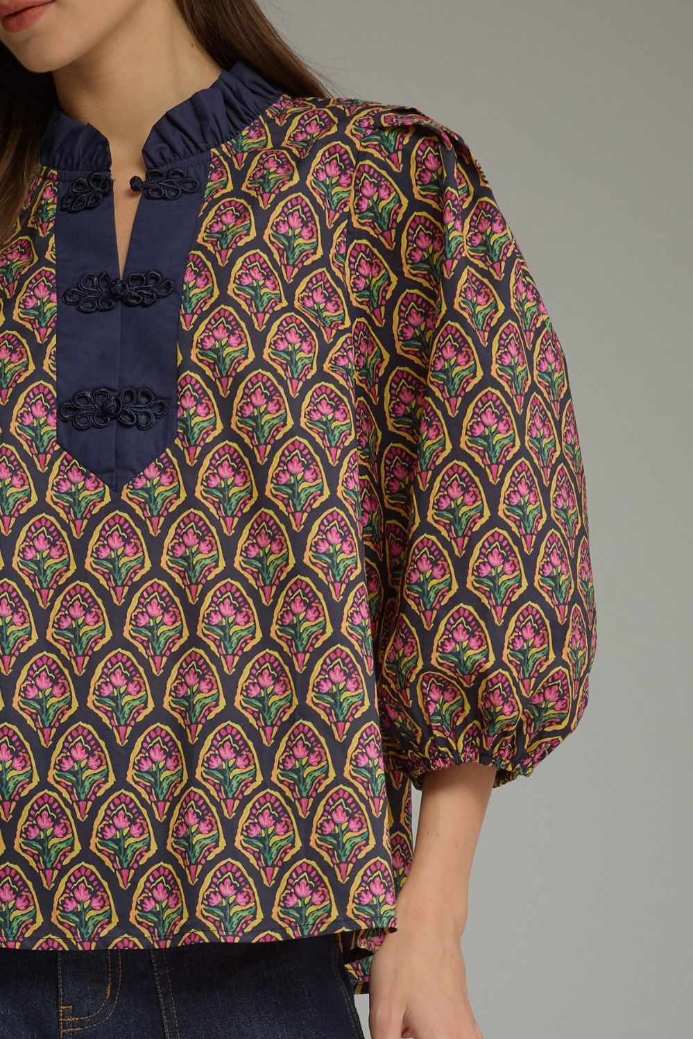 A close-up of a woman wearing a patterned blouse with a navy blue ruffled collar, three decorative black buttons, and voluminous sleeves. The blouse features a repeating pink, green, and gold floral arch pattern on a dark background.