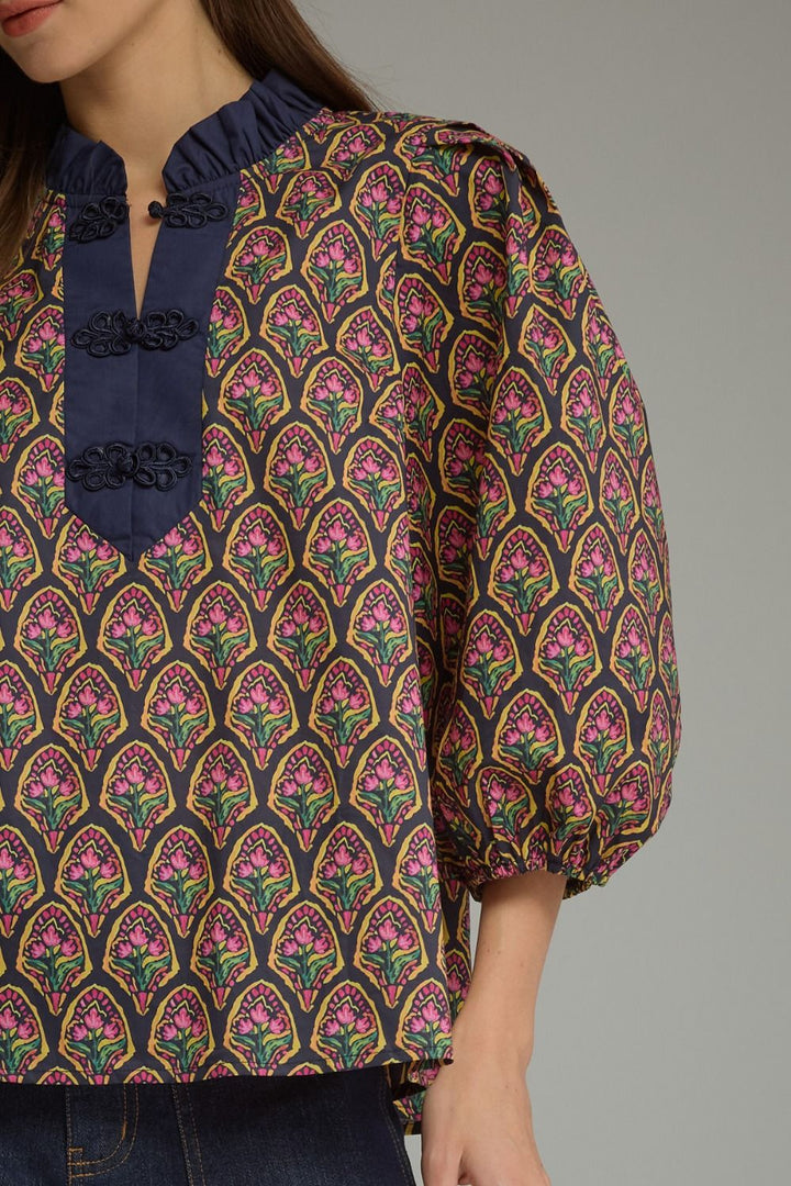 A close-up of a woman wearing a patterned blouse with a navy blue ruffled collar, three decorative black buttons, and voluminous sleeves. The blouse features a repeating pink, green, and gold floral arch pattern on a dark background.