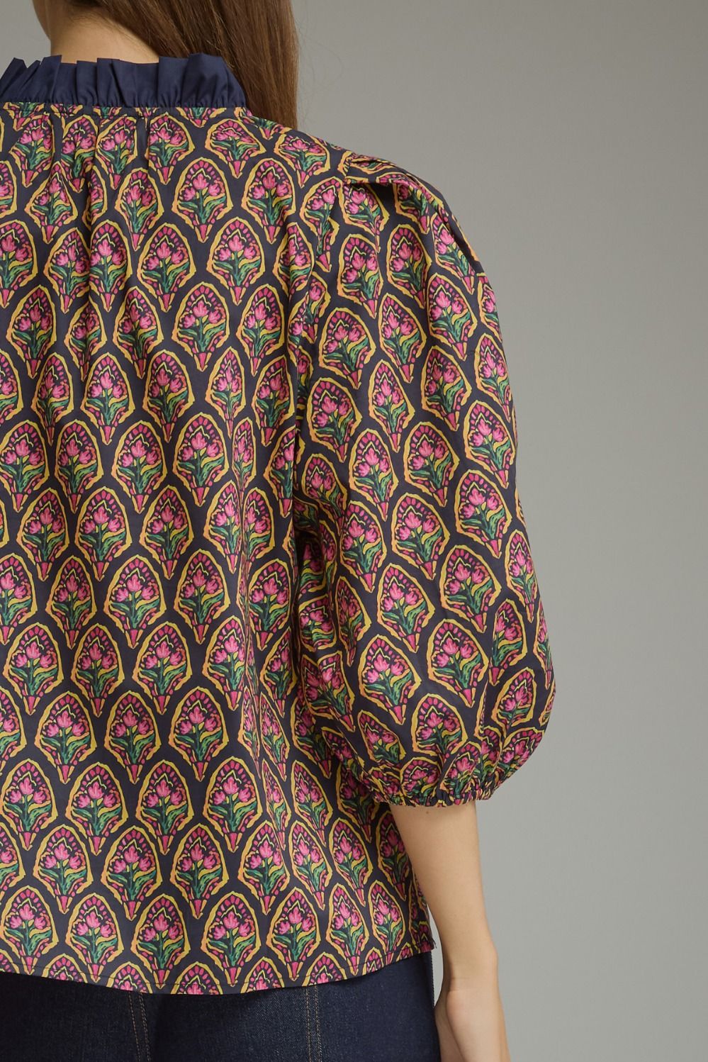 A close-up of a woman wearing a patterned blouse with puff sleeves and a dark ruffled neckline. The blouse features an intricate repeating arch pattern in pink, green, yellow, and black.