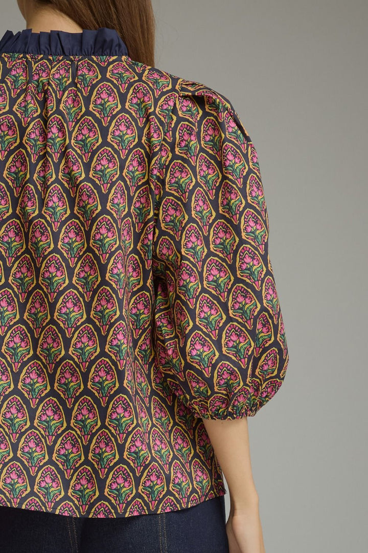 A close-up of a woman wearing a patterned blouse with puff sleeves and a dark ruffled neckline. The blouse features an intricate repeating arch pattern in pink, green, yellow, and black.