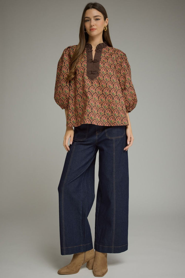 A woman modeling a brown patterned blouse with a high collar and wide sleeves, paired with dark wide-leg jeans and tan ankle boots, against a neutral gray studio background.