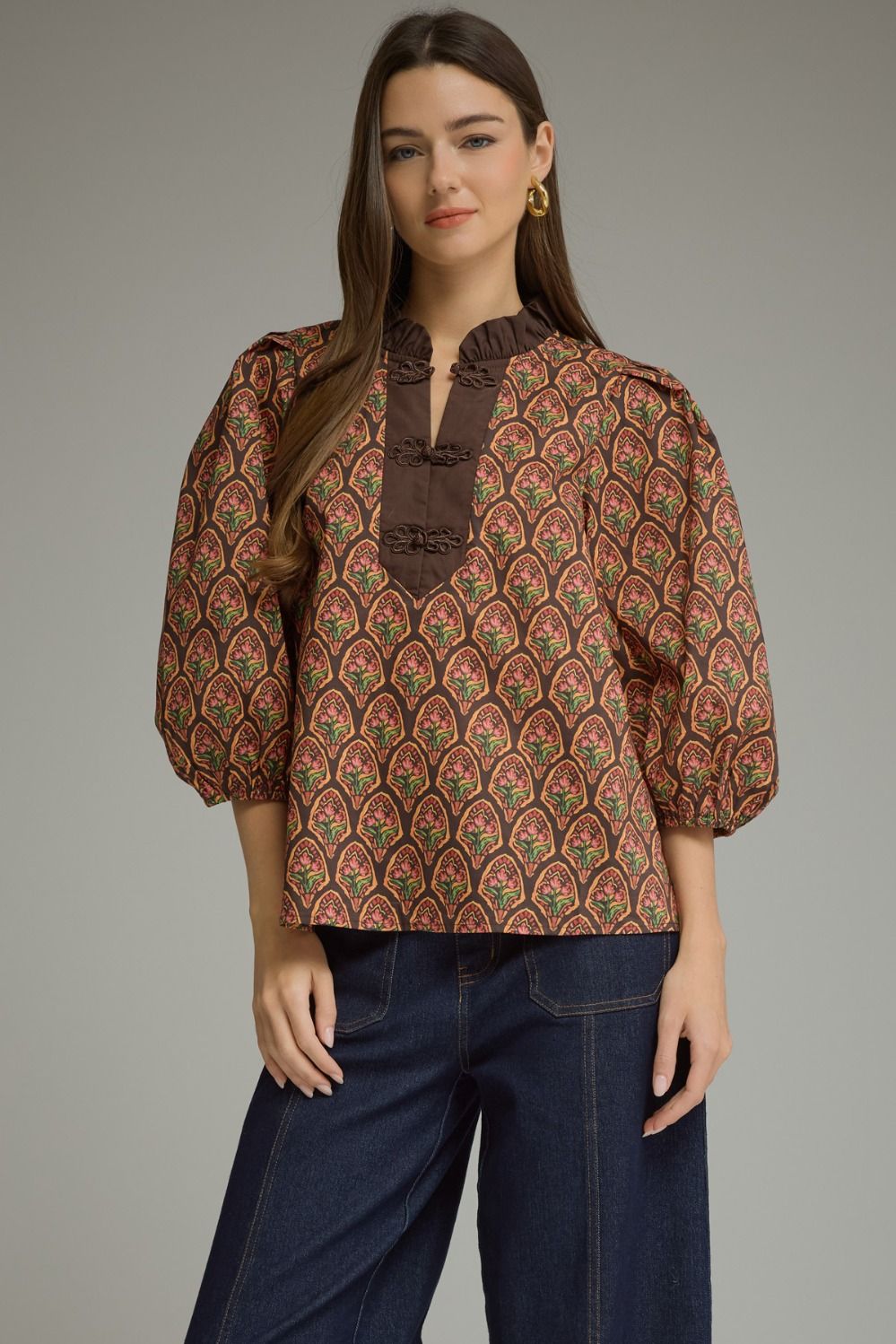 A woman modeling a brown patterned blouse with puff sleeves and a dark brown ruffled mock neck, paired with dark blue jeans, against a solid gray studio background.