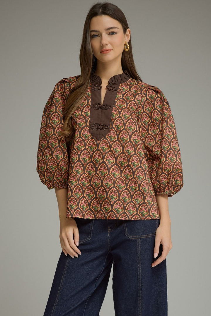 A woman modeling a brown patterned blouse with puff sleeves and a dark brown ruffled mock neck, paired with dark blue jeans, against a solid gray studio background.