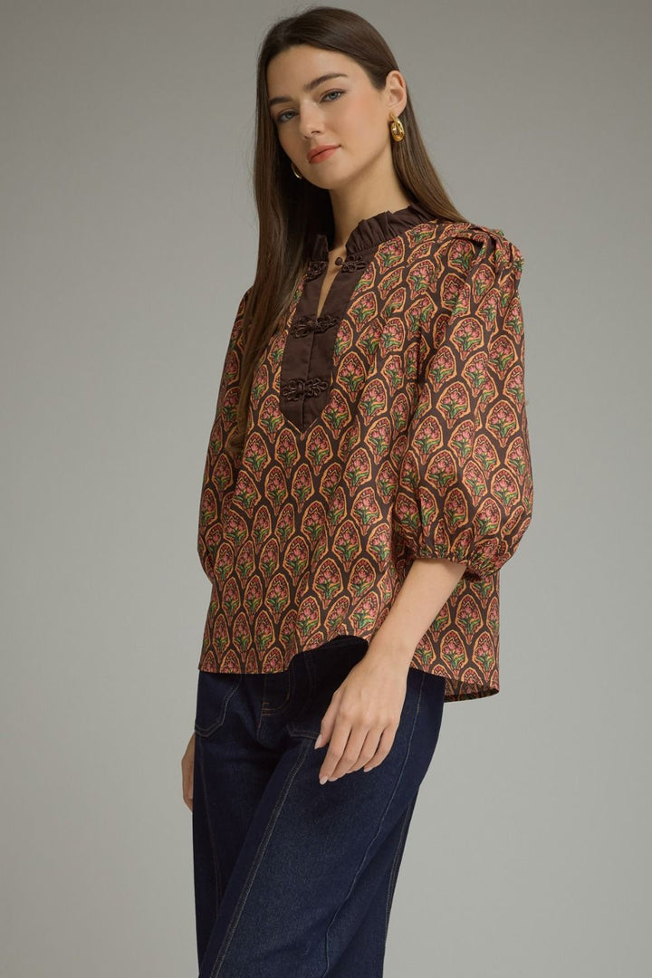 A woman modeling a brown patterned blouse with puff sleeves and a dark brown ruffled neckline, paired with dark blue jeans, against a solid gray studio background.