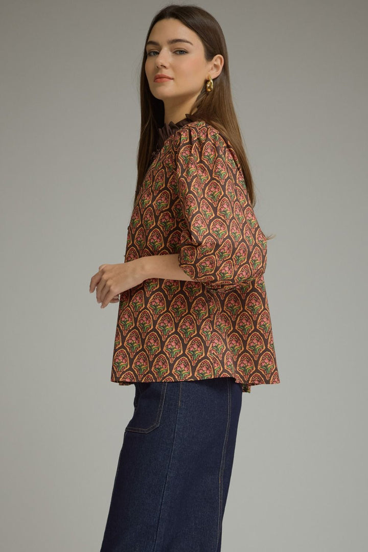 A woman wearing a brown patterned blouse with red, green, and gold paisley-like motifs, paired with dark blue jeans, photographed against a plain gray studio background.