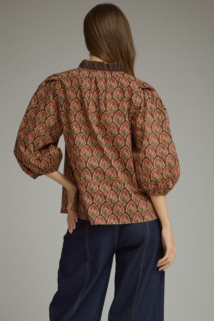 A woman shown from the back wearing a brown patterned blouse with puff sleeves and dark blue jeans, photographed against a plain gray studio background.