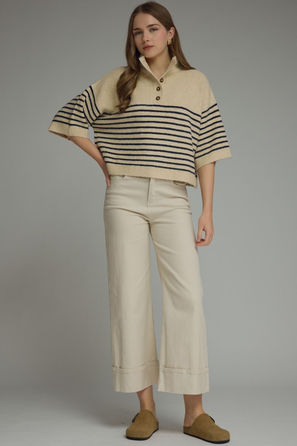 A woman modeling a cropped knit polo sweater with beige and black horizontal stripes, paired with cream cropped trousers and tan slip-on shoes, against a solid gray studio background.