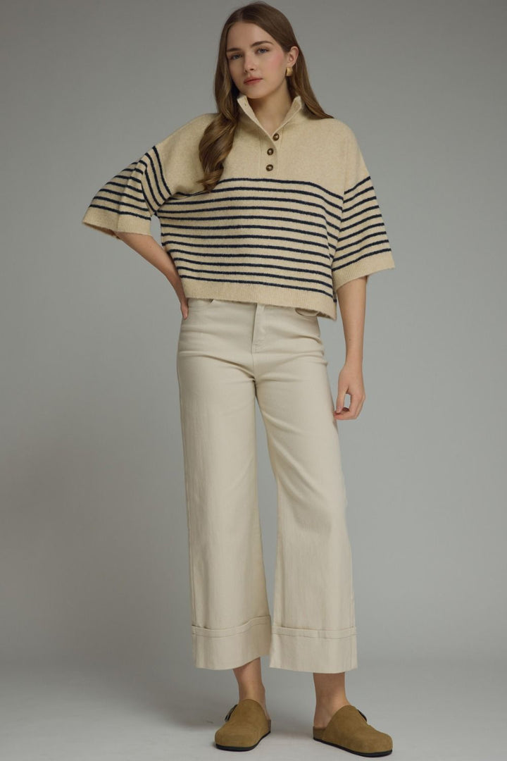 A woman modeling a cropped knit polo sweater with beige and black horizontal stripes, paired with cream cropped trousers and tan slip-on shoes, against a solid gray studio background.