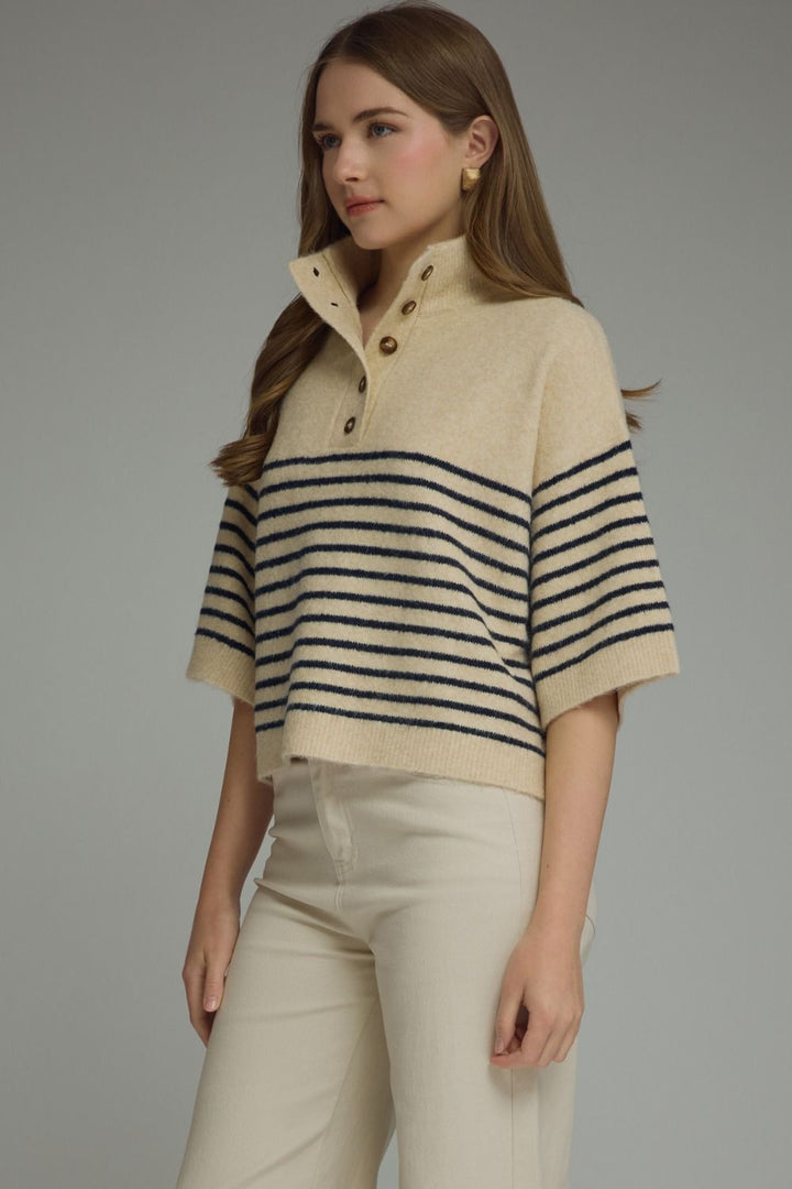 A woman wearing a cropped knit polo sweater with horizontal navy stripes on a cream base, paired with cream pants, against a solid gray studio background.