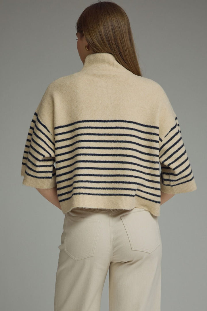 A woman shown from the back wearing a cream knit sweater with navy horizontal stripes across the back and sleeves, paired with cream pants, against a solid gray studio background.
