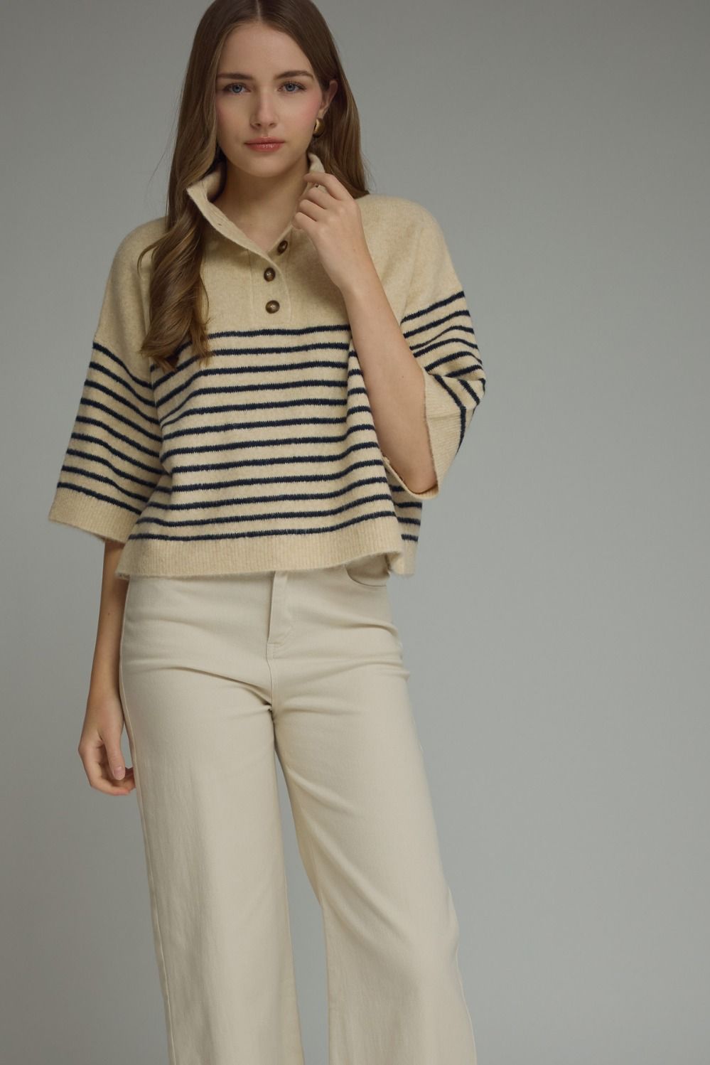 A woman modeling a cropped knit polo sweater with horizontal navy and beige stripes, paired with light beige wide-leg trousers, against a solid gray studio background.