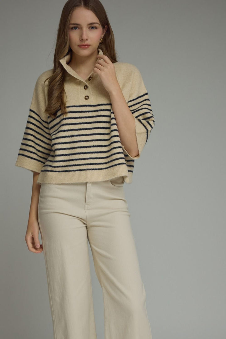 A woman modeling a cropped knit polo sweater with horizontal navy and beige stripes, paired with light beige wide-leg trousers, against a solid gray studio background.