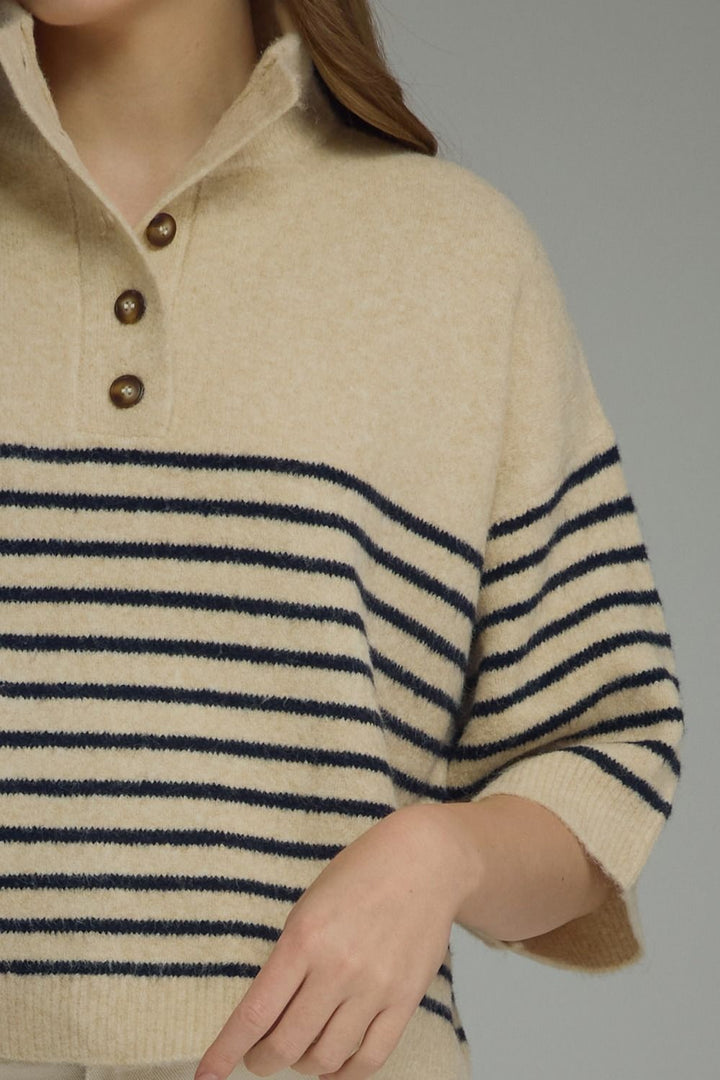 Close-up of a person wearing a cream knit sweater with navy horizontal stripes across the chest and sleeves, plus three brown buttons at the collar, against a plain gray background.