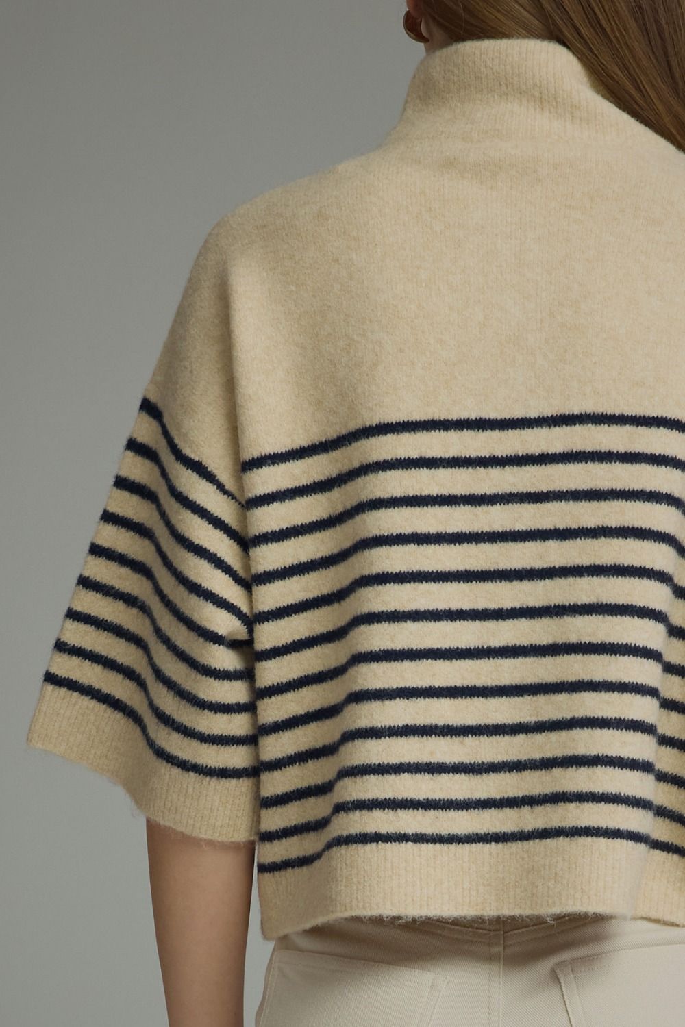 Close-up back view of a person wearing a cream knit turtleneck sweater with navy horizontal stripes across the shoulders and upper back, paired with light beige pants.