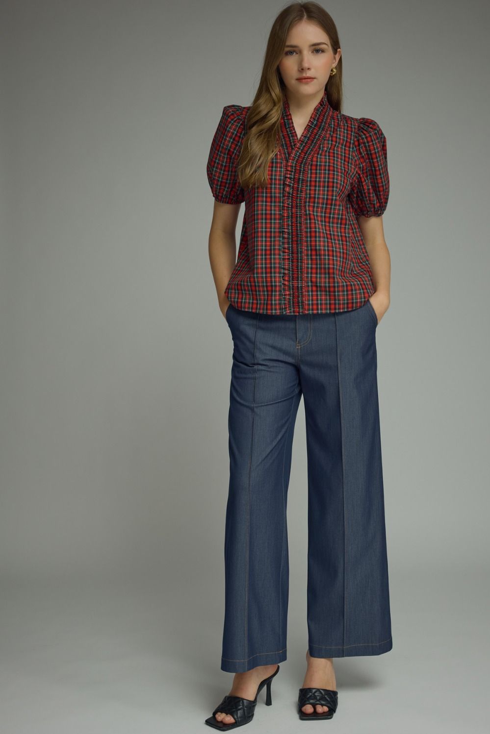 A woman modeling a short-sleeve plaid button-up blouse with puff sleeves in red, blue, and black, paired with wide-leg blue jeans and black heeled sandals, against a solid gray studio background.
