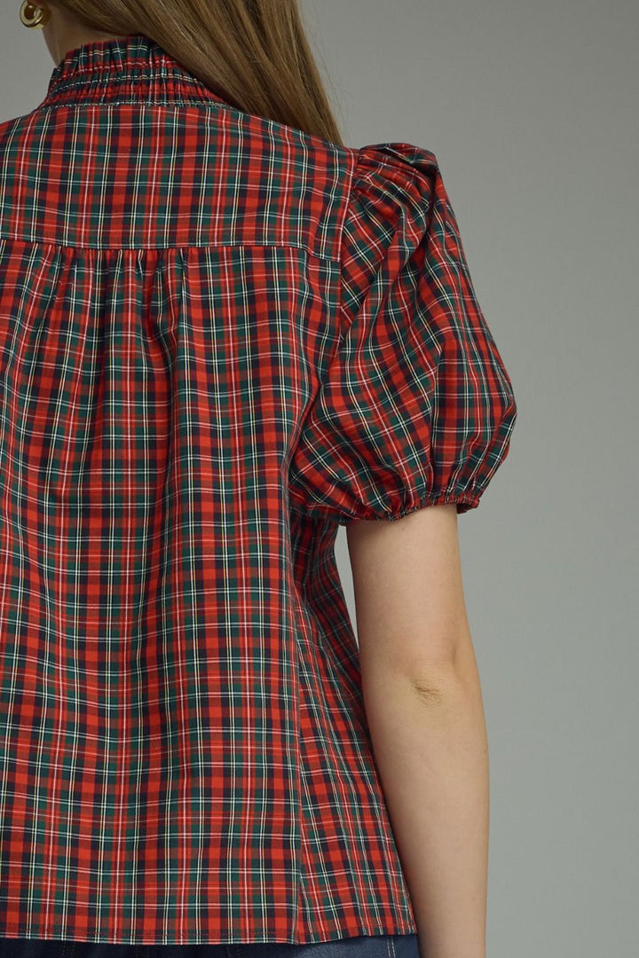 A close-up back view of a model wearing a red and green plaid short-sleeve blouse with puff sleeves, set against a plain gray background.