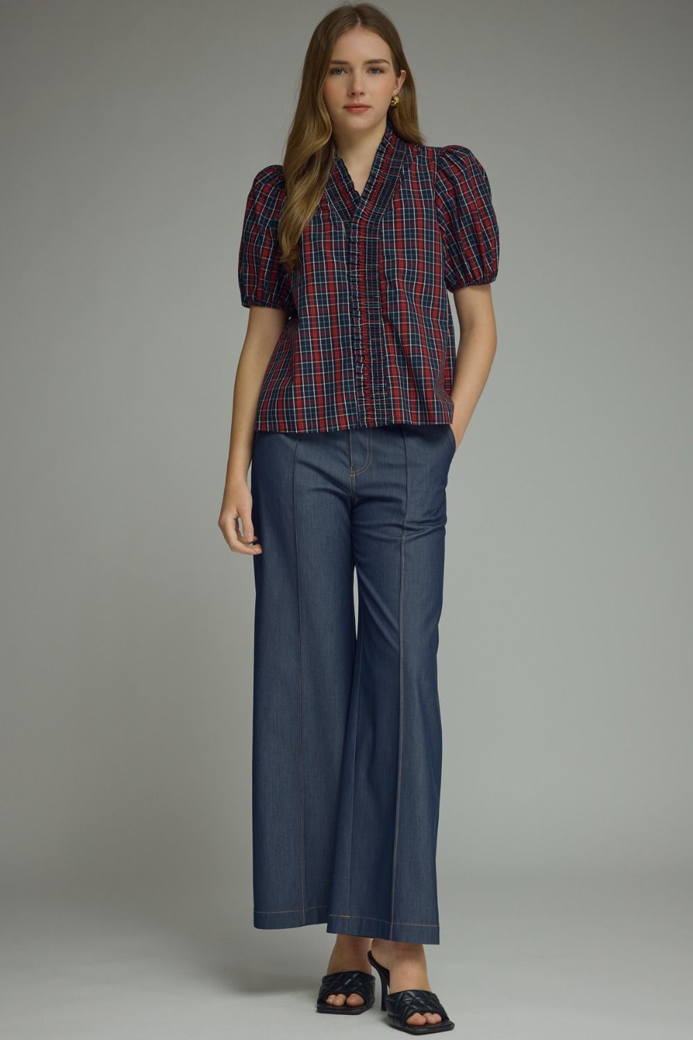 A woman modeling a short-sleeve plaid button-up shirt in navy, red, and white, paired with dark blue wide-leg trousers and black slide sandals, against a neutral gray studio background.