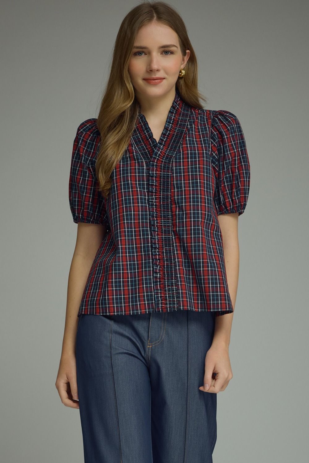 A woman modeling a short-sleeve plaid blouse with puff sleeves and a gathered front, paired with dark blue pants, against a plain gray studio background.