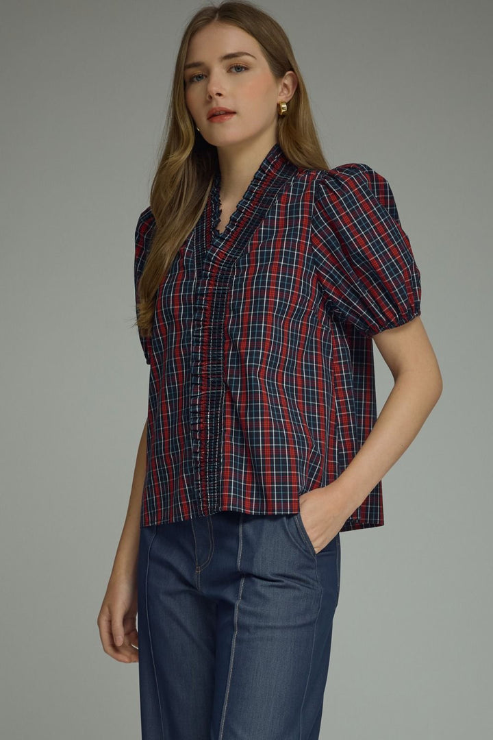 A woman modeling a short-sleeve plaid blouse with puff sleeves and a wrap-style front, paired with blue jeans, against a plain gray studio background.