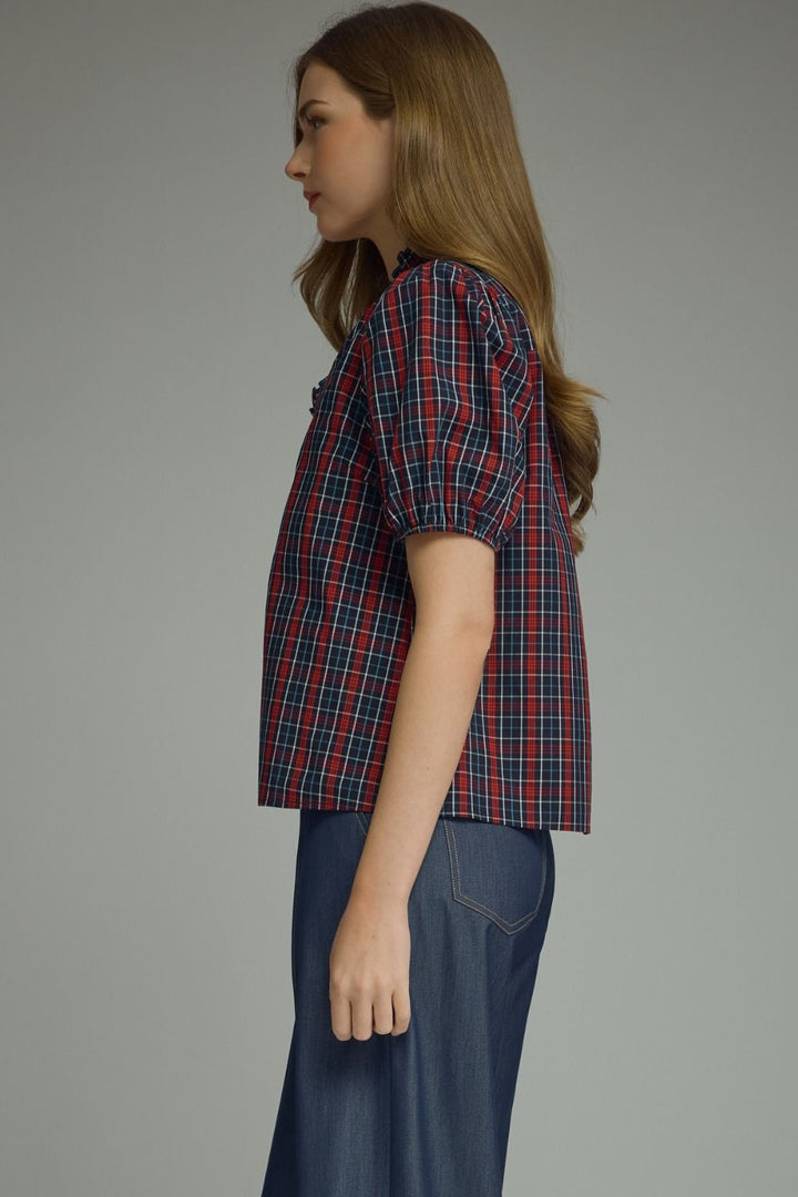 A side-profile view of a young woman wearing a short-sleeve plaid button-up shirt in red, navy, and white, paired with dark blue jeans, against a solid gray studio background.