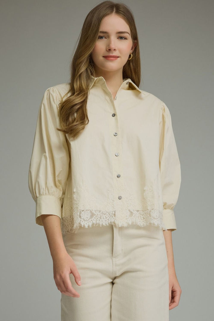 A woman modeling a cream-colored long-sleeve button-up blouse with lace trim at the hem and matching cream pants, photographed against a solid gray studio background.