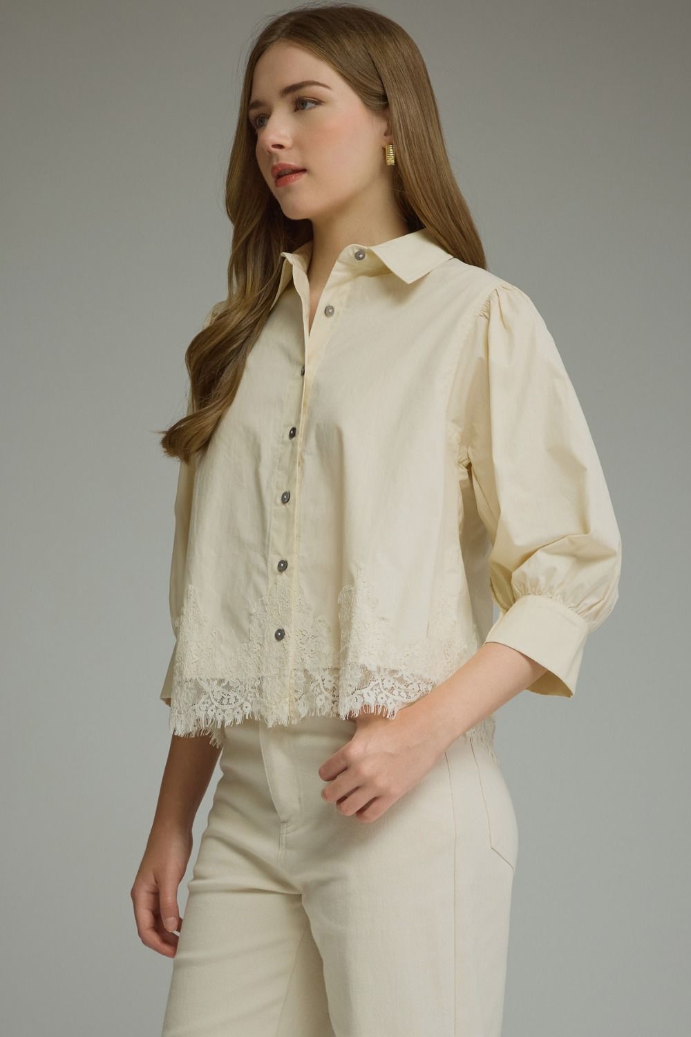 A woman wearing a cream-colored button-up blouse with lace trim at the hem and matching cream trousers, photographed against a neutral gray studio background.