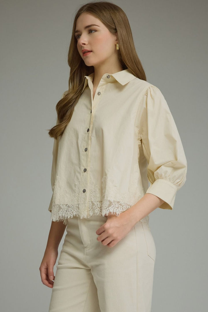 A woman wearing a cream-colored button-up blouse with lace trim at the hem and matching cream trousers, photographed against a neutral gray studio background.