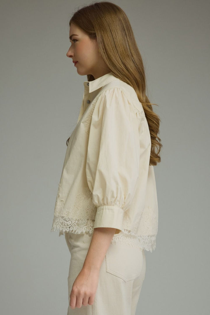 A woman shown in side profile wearing a cream-colored short-sleeve blouse with lace trim at the hem and cuffs, paired with matching cream trousers, against a solid gray studio background.