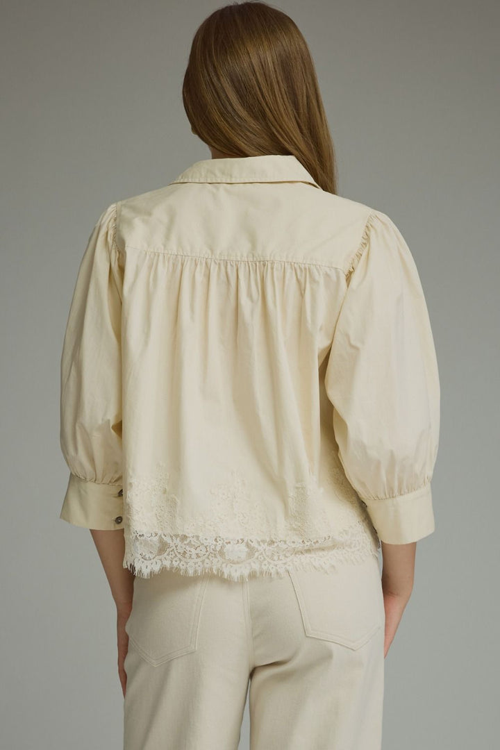 A woman shown from the back wearing a cream-colored blouse with puffed sleeves, gathered detailing, and a lace-trimmed hem, paired with matching cream trousers, against a solid gray studio background.
