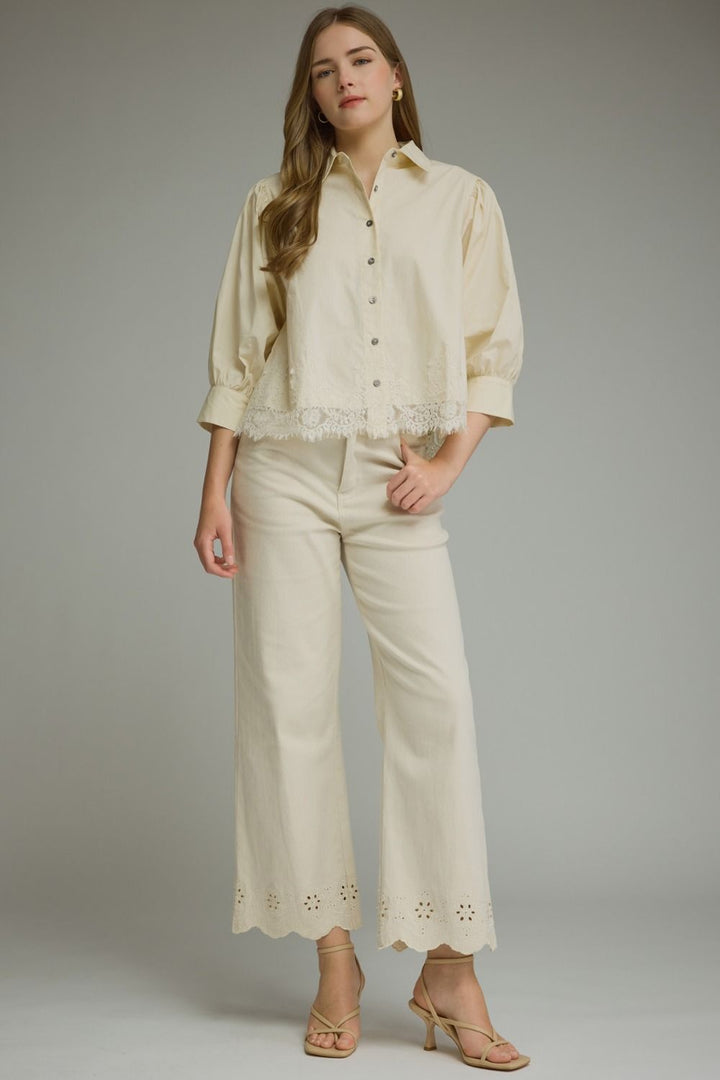 A woman modeling a cream-colored two-piece set consisting of a long-sleeve button-up blouse with eyelet lace trim and matching wide-leg cropped pants with eyelet lace detailing at the hem, paired with beige strappy heels against a neutral gray studio background.