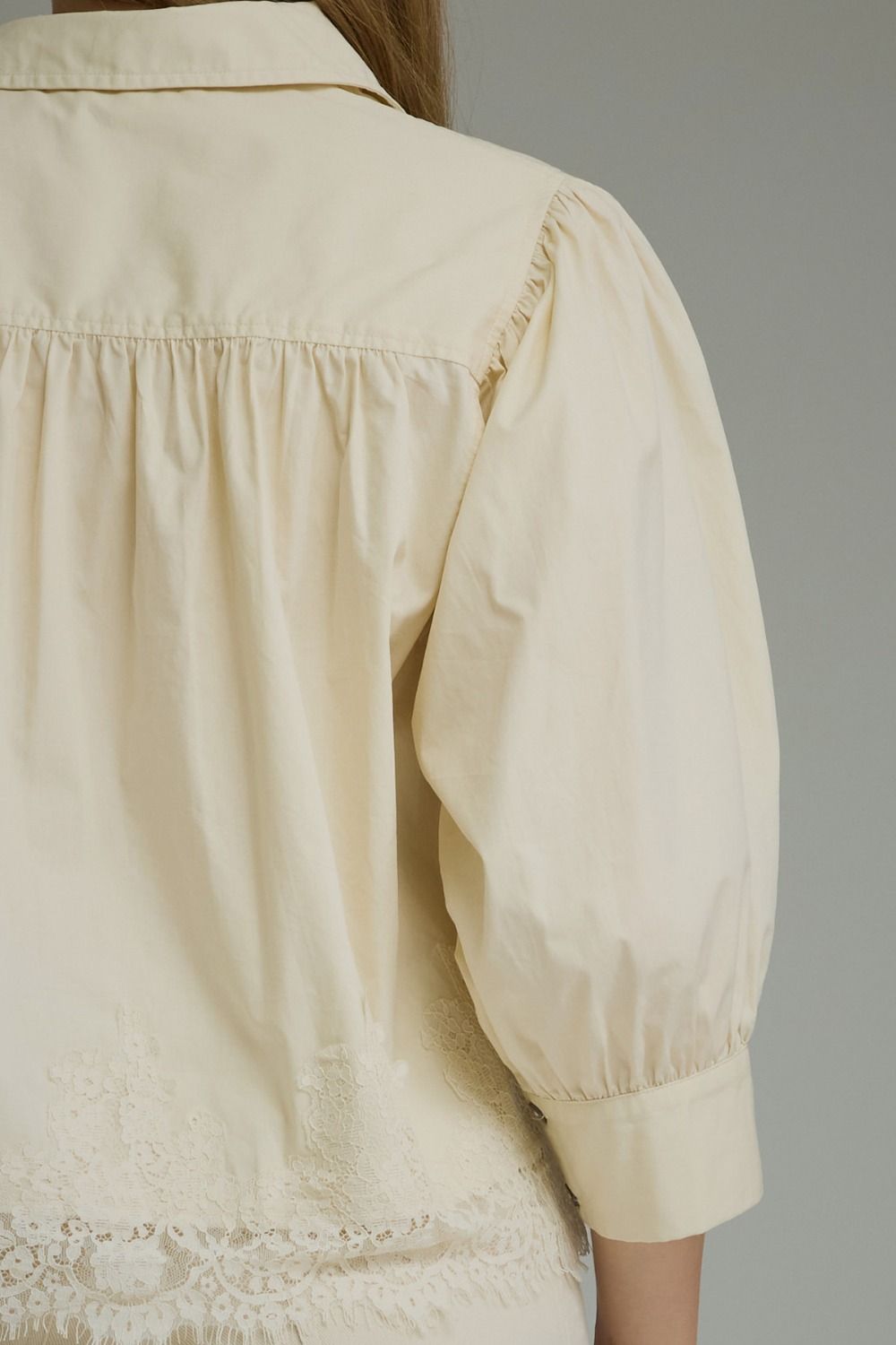 Close-up back view of a cream-colored blouse with gathered shoulders, puffed sleeves, and lace trim at the hem, shown on a model against a neutral gray background.
