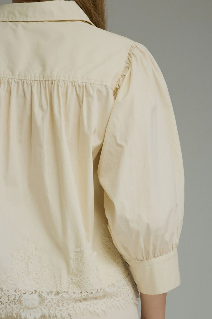 Close-up back view of a cream-colored blouse with gathered shoulders, puffed sleeves, and lace trim at the hem, shown on a model against a neutral gray background.