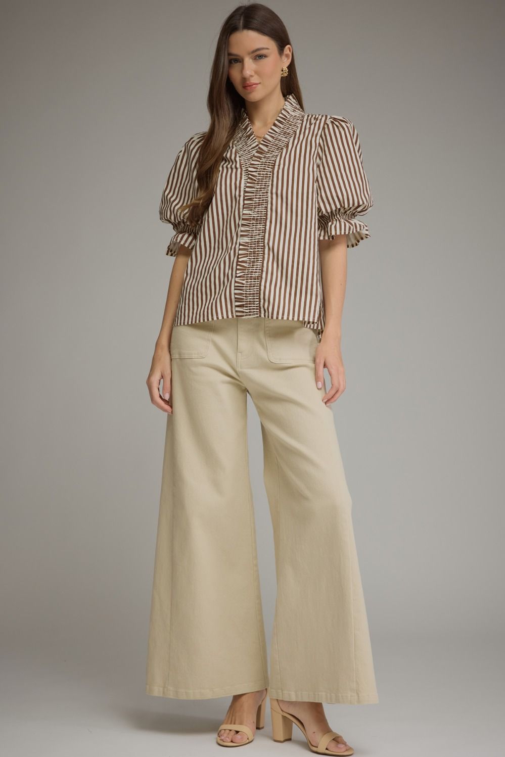 A woman modeling a brown and beige vertically striped blouse with ruffled trim and puffed sleeves, paired with wide-leg beige trousers and nude heels, against a solid gray studio background.