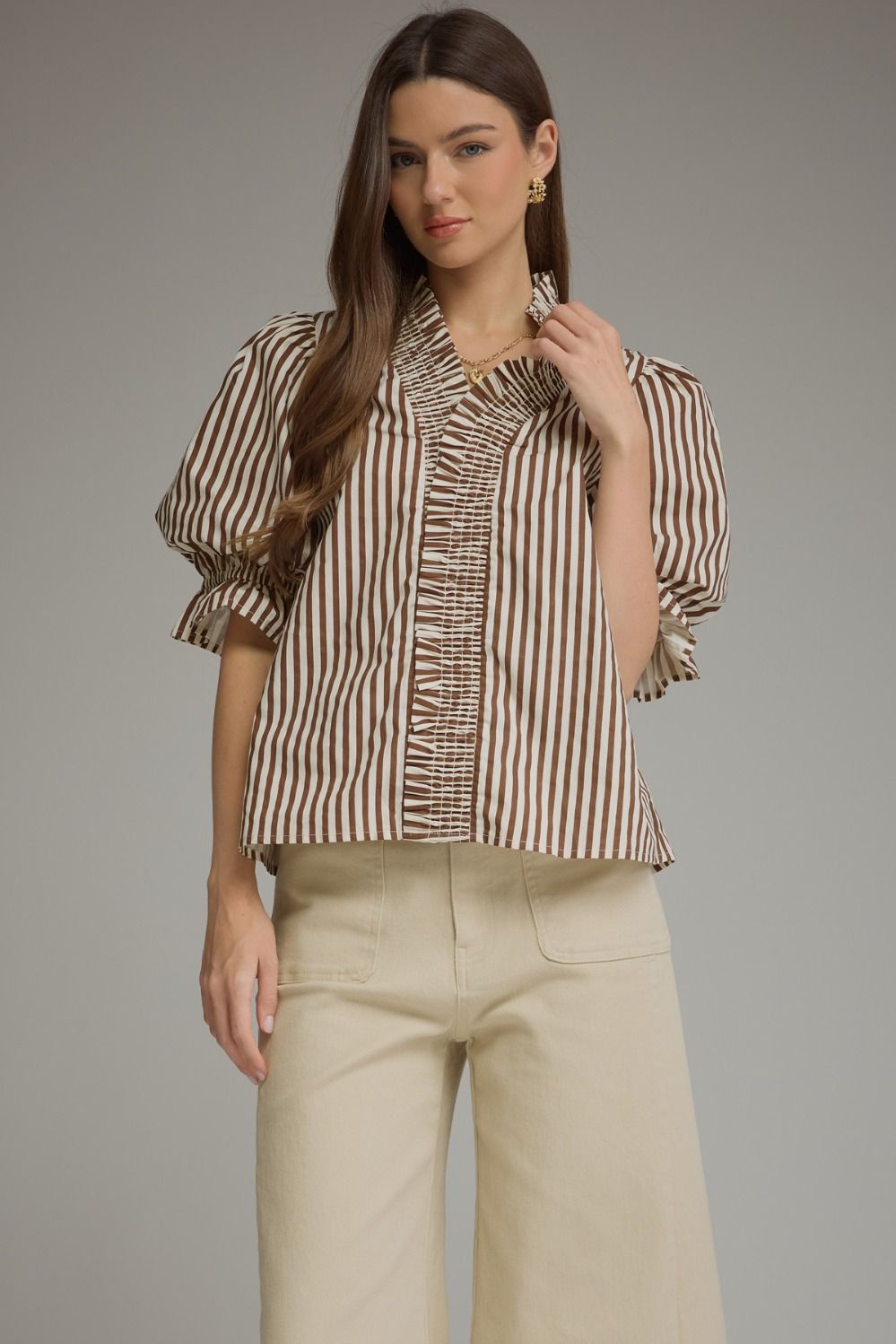 A woman modeling a brown and beige vertically striped blouse with puff sleeves and a ruffled front, paired with light beige wide-leg pants, against a solid gray studio background.