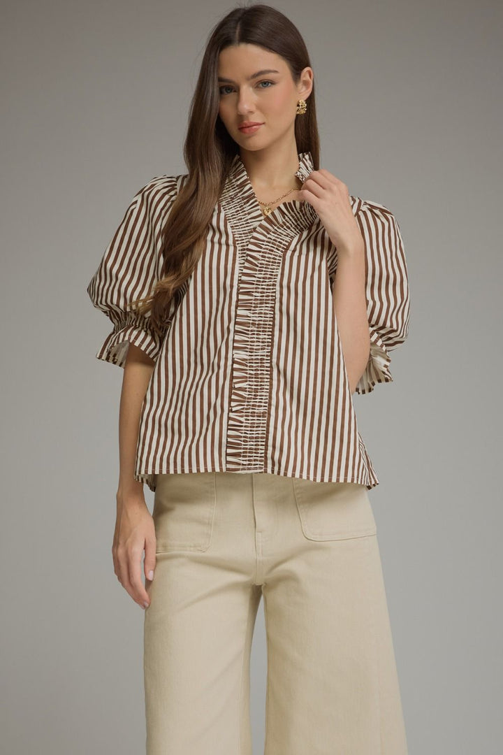 A woman modeling a brown and beige vertically striped blouse with puff sleeves and a ruffled front, paired with light beige wide-leg pants, against a solid gray studio background.