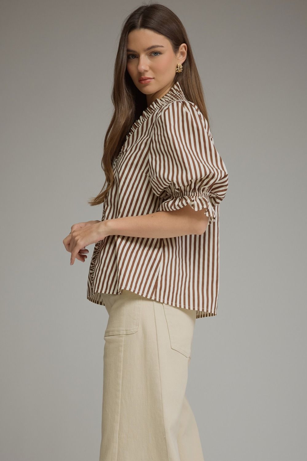 A woman modeling a brown and beige vertically striped blouse with ruffled sleeves, paired with cream-colored trousers, against a solid gray studio background.