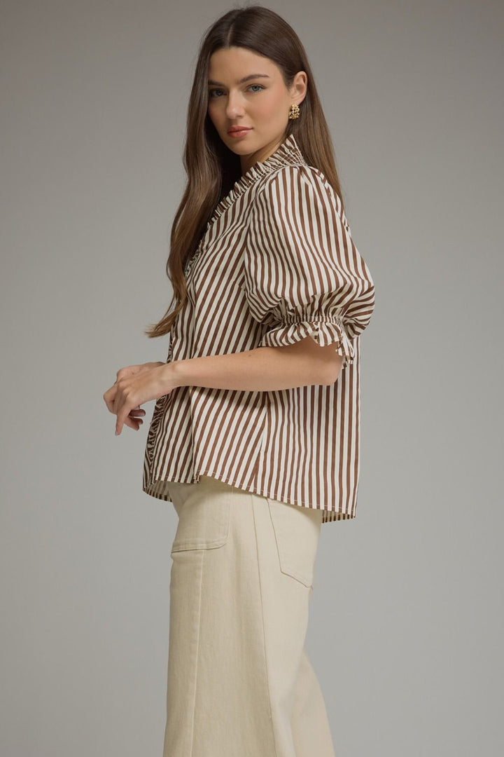 A woman modeling a brown and beige vertically striped blouse with ruffled sleeves, paired with cream-colored trousers, against a solid gray studio background.
