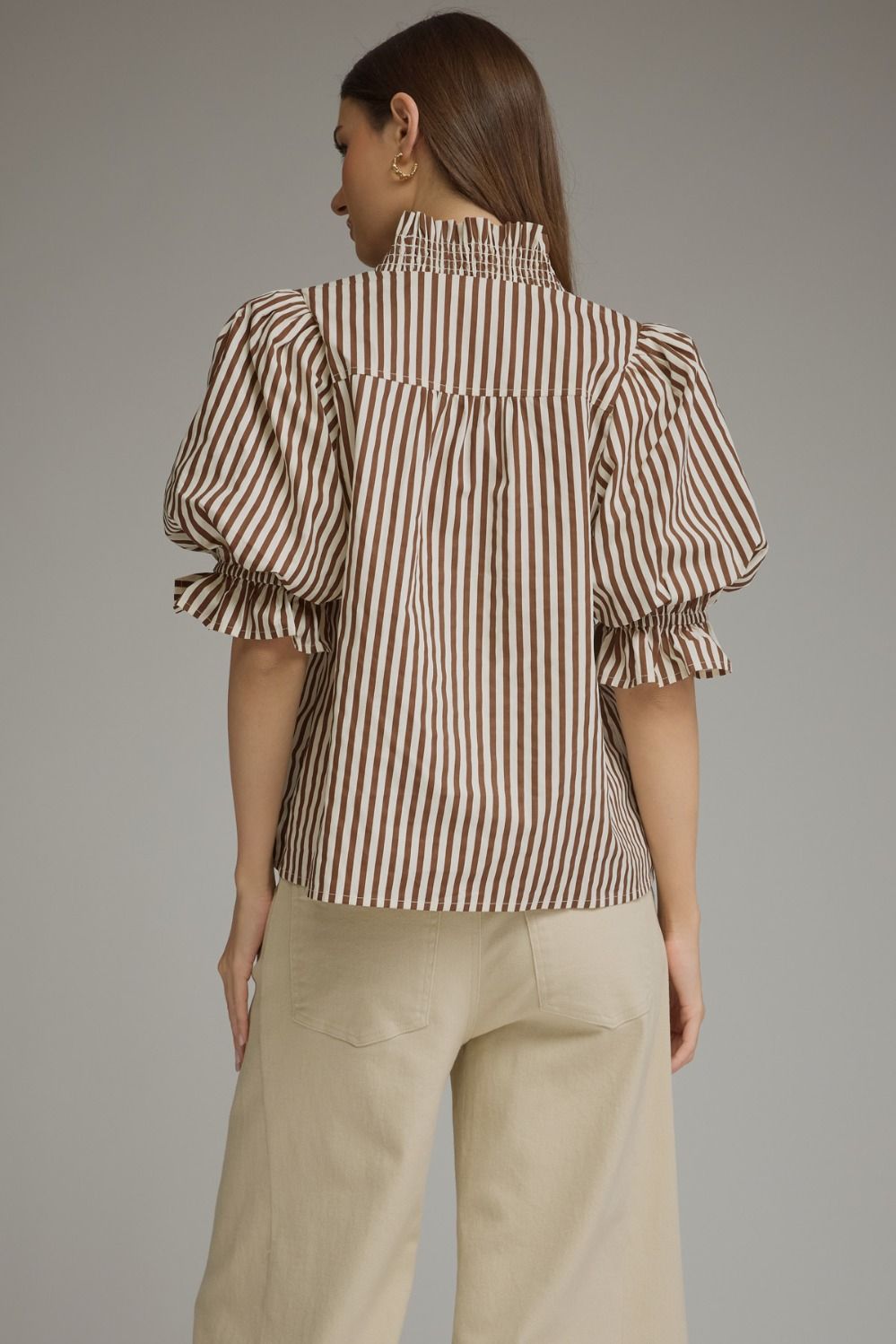 A woman shown from the back wearing a brown and beige vertically striped blouse with puffed ruffled sleeves and a smocked high neckline, paired with beige pants, against a solid gray studio background.